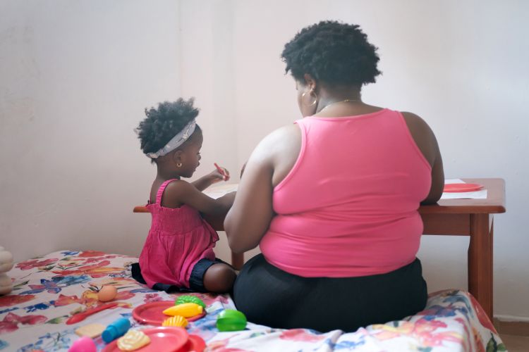 A woman and a young girl, both wearing pink tops, sit on a bed with a floral blanket, facing away from the camera. The girl is playing with colorful toy food and utensils on the bed near a small table.