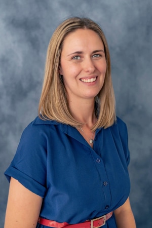 A woman with straight, shoulder-length blonde hair is wearing a blue short-sleeve button-up shirt and a red belt. She is smiling and posing in front of a mottled gray background.