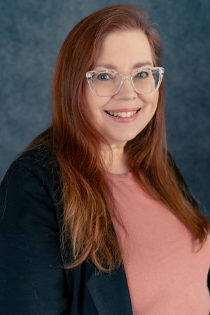 A woman with long reddish-brown hair wearing clear glasses, a black blazer, and a pink top smiles in front of a blue-gray textured background.