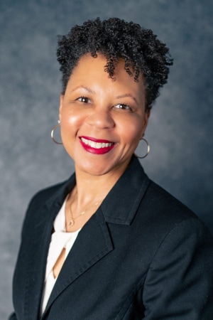 A woman with short curly hair, wearing hoop earrings, a black blazer, and a white top, smiles against a dark, textured background.