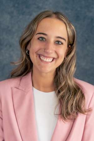 A young woman with long, wavy, light brown hair smiles at the camera. She is wearing a pink blazer over a white top and has a nose ring. The background is a textured blue-gray.