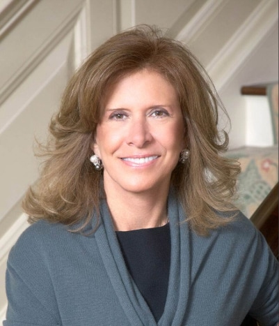 A woman with shoulder-length light brown hair, wearing a gray shawl over a black top and pearl earrings, smiles while seated indoors against a background of white paneled stairs and a patterned chair.
