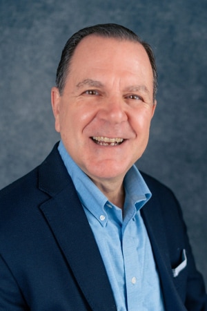A middle-aged man in a blue blazer and light blue shirt smiles at the camera against a textured blue-gray background.