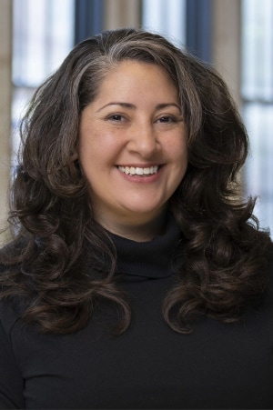 A woman with long, wavy dark hair and streaks of gray smiles at the camera. She is wearing a black turtleneck sweater and stands in front of a blurred indoor background with large windows.