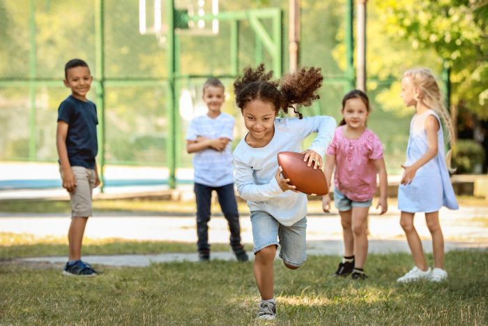 Five children play outside on grass. One girl in front runs while holding a football, smiling, as the other children watch and follow her. Trees and a sports court are visible in the background on a sunny day.