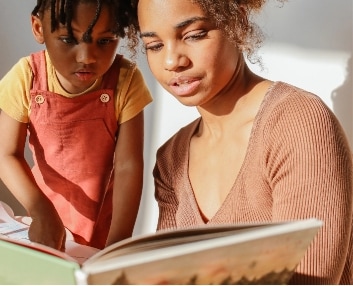 An adult and a child sit together reading a book in bright natural light. The adult holds the book open while the child, wearing a red pinafore, looks at the pages with interest.