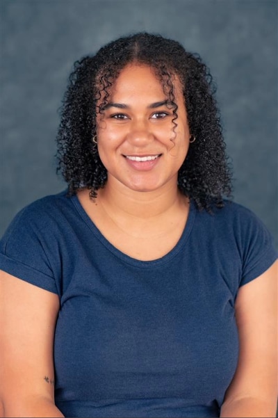 A woman with curly dark hair and a navy blue t-shirt smiles while sitting in front of a plain, dark background.