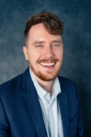 A man with short curly brown hair and a beard smiles at the camera. He is wearing a dark blue blazer over a light-colored shirt, set against a dark, textured background.