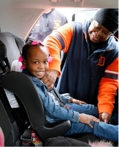 A smiling young girl sits in a car seat while an adult, wearing a jacket and hat, fastens her seatbelt inside a vehicle.