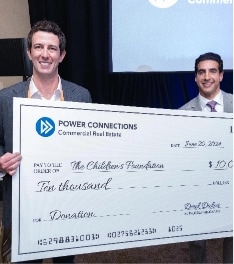 Two men in suits smile while holding an oversized check for $10,000 made out to The Children's Foundation from Power Connections Commercial Real Estate. The date on the check is June 20, 2023.