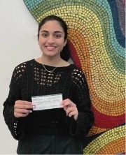 A woman in a black sweater smiles while holding a check. She stands in front of a colorful, abstract mosaic wall.