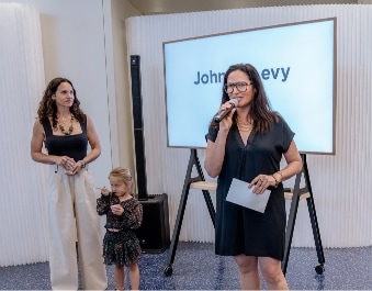 A woman in a black dress speaks into a microphone while holding a paper. Another woman and a young girl stand nearby. Behind them is a large screen displaying partially visible text, "John...evy.