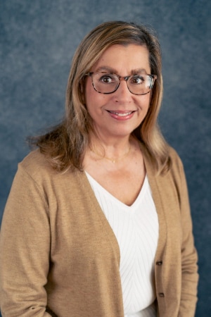 A woman with long light brown hair, wearing glasses, a tan cardigan, and a white top, smiles softly while standing in front of a blue textured background.