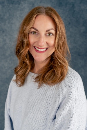 A woman with wavy light brown hair, wearing a light gray sweater, smiles at the camera against a textured blue background.