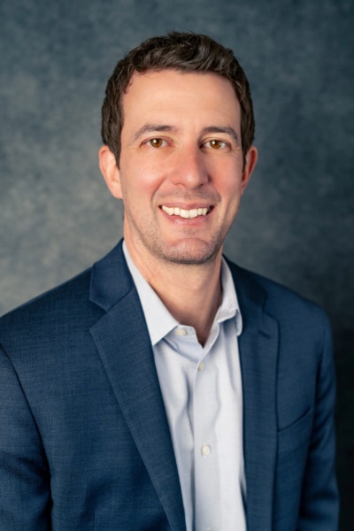 A man with short dark hair, wearing a dark blue suit jacket and a light blue shirt, smiles at the camera against a mottled gray background.