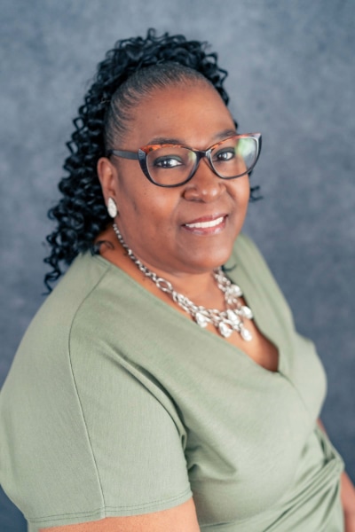 A woman with glasses and curly hair pulled back smiles at the camera. She is wearing a light green top, a statement necklace, and earrings, with a grey textured background behind her.