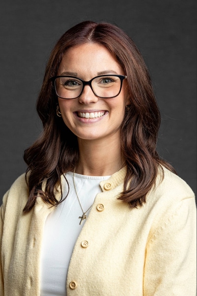 A woman with shoulder-length brown hair and glasses smiles at the camera. She is wearing a cream-colored cardigan over a white top and a necklace with a cross pendant. The background is plain and dark.
