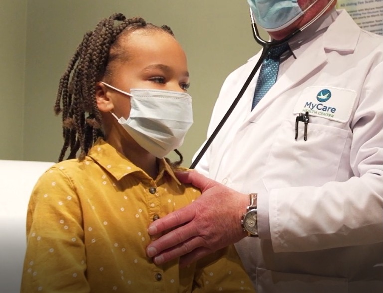 A child wearing a mask sits on an exam table while a doctor in a white coat and mask uses a stethoscope to listen to the child's chest.