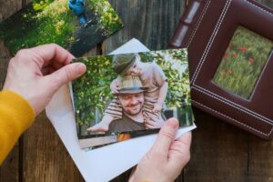A person holds a photo of a man with a child on his shoulders, both smiling outdoors. An envelope, another photo, and a leather photo album are on a rustic wooden table.