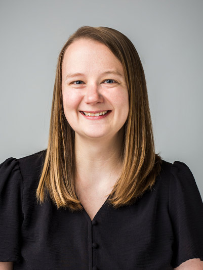 A woman with straight, shoulder-length light brown hair smiles at the camera. She is wearing a black blouse and is posed against a plain light gray background.