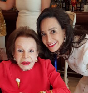 Two women pose closely together and smile. The older woman, in a jeweled red top and gold jewelry, shares a warm moment with the younger woman in white. Their bond reflects the spirit of legacy giving. A bar with bottles is visible in the background.