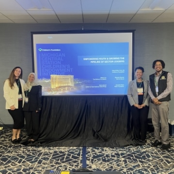 Four people stand smiling in front of a presentation screen displaying a slide titled “Empowering Youth & Growing the Pipeline of Sector Leaders” at a conference. The background features patterned carpet and gray walls.