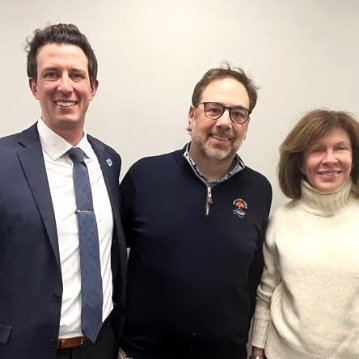 Three adults stand together, smiling at the camera. The man on the left wears a dark suit and tie, the man in the center wears glasses and a navy pullover, and the woman on the right wears a white turtleneck sweater.