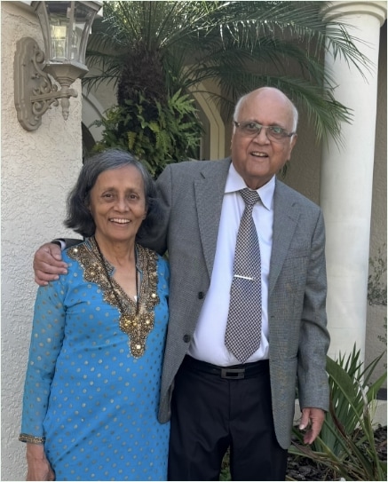 An older woman in a blue dress and an older man in a suit and tie stand smiling together outside a building with columns and greenery in the background.