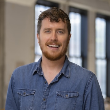 A man with short, curly brown hair and a beard, wearing a blue denim shirt, smiles while standing indoors in front of large windows with blurred light from outside.