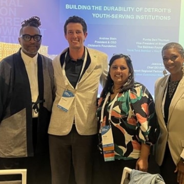 Four people stand together and smile at a conference in front of a screen that reads "Building the Durability of Detroit's Youth-Serving Institutions." They wear business or business casual attire and conference badges.