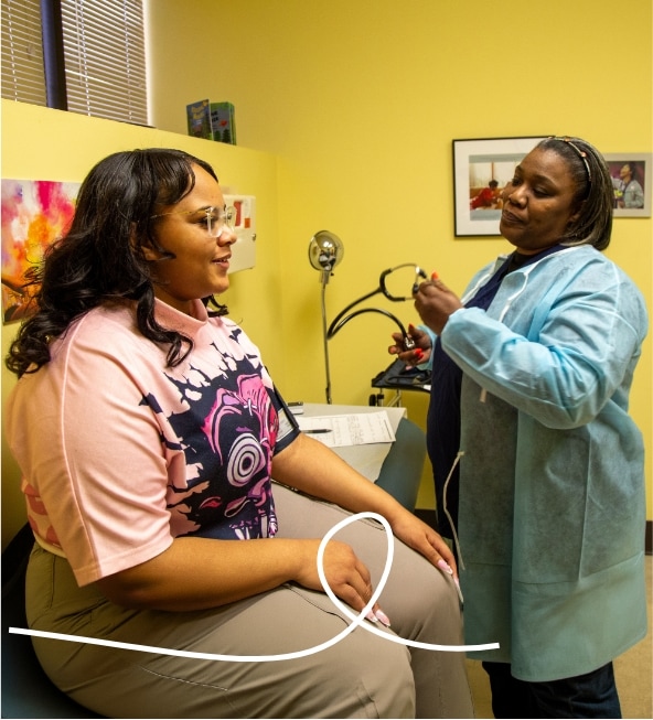 A healthcare worker wearing a blue gown and gloves prepares a syringe while a seated woman in a pink shirt with a bold graphic print looks on in a medical examination room with yellow walls.