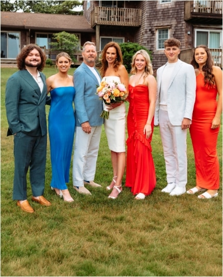 A group of six people pose outside on grass, dressed in formal summer attire. The woman in the center holds a bouquet, wearing a white dress, surrounded by others in colorful suits and dresses, with a house in the background.
