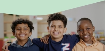 Three smiling children stand close together indoors, each with an arm around the other's shoulder.