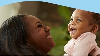 Smiling woman holding a happy baby who is wearing a pink outfit. Both are looking at each other with joy, and the background is softly blurred with some greenery visible.