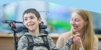 A smiling boy in a wheelchair holds hands with a woman who is also smiling at him. The background is softly blurred, focusing on their interaction.