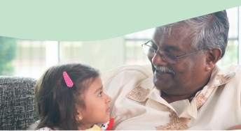 An older man with gray hair and glasses smiles warmly at a young girl with a pink hair clip as they sit together on a couch.