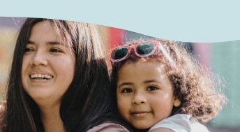 A woman with long dark hair smiles while a young girl with curly hair and pink sunglasses on her head hugs her from behind.