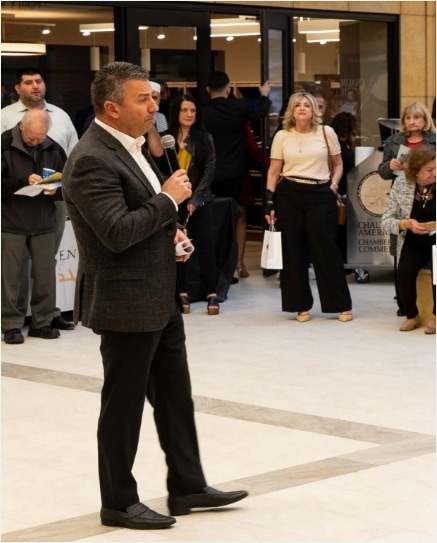 A man in a suit speaks into a microphone in a mall or public indoor space while several people stand and listen in the background.