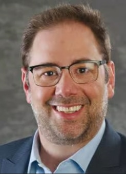 A man with light skin, short brown hair, and a beard is smiling. He is wearing glasses, a blue shirt, and a dark blazer, with a blurred gray background behind him.