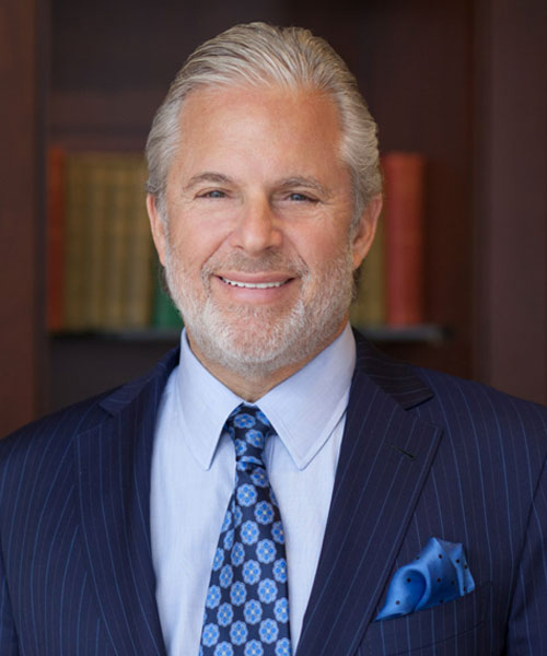Smiling middle-aged man with gray hair and beard, wearing a blue pinstripe suit, light blue shirt, and patterned tie, standing in front of a bookshelf.