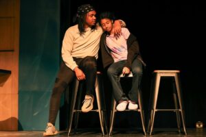 Two people sit on tall stools on a stage. One person has their arm around the other's shoulders, comforting them. Warm light highlights their faces against a dark background.
