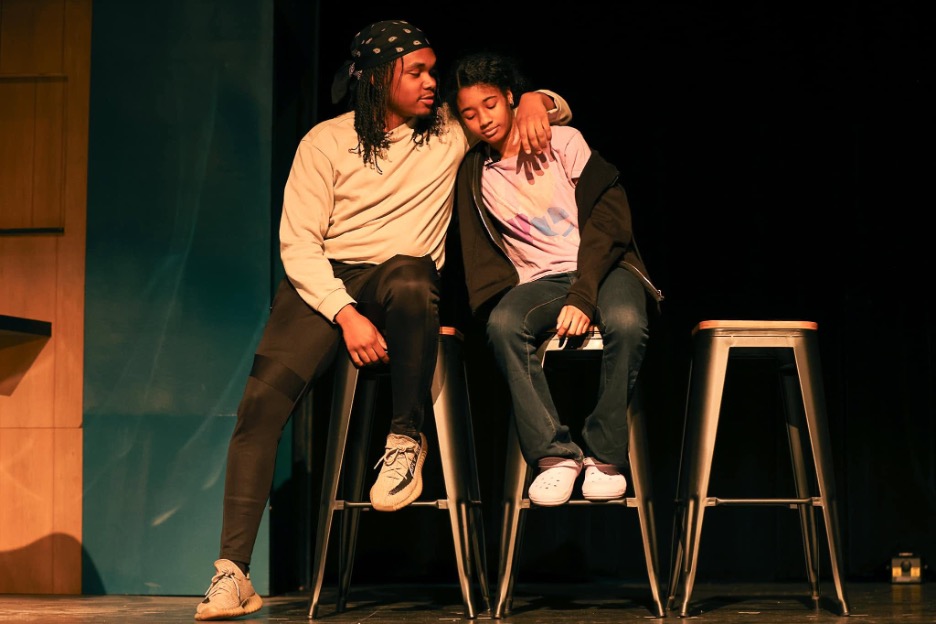 Two people sit on tall stools on a stage. One person has their arm around the other's shoulders, comforting them. Warm light highlights their faces against a dark background.