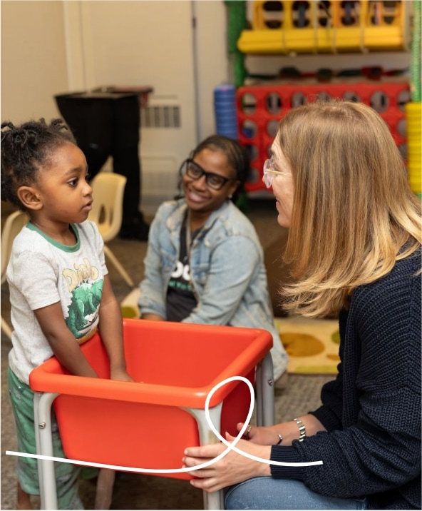 A young child stands at a red sensory table while a woman with glasses kneels and talks to them. Another adult sits in the background, smiling. The setting appears to be a classroom or play area.