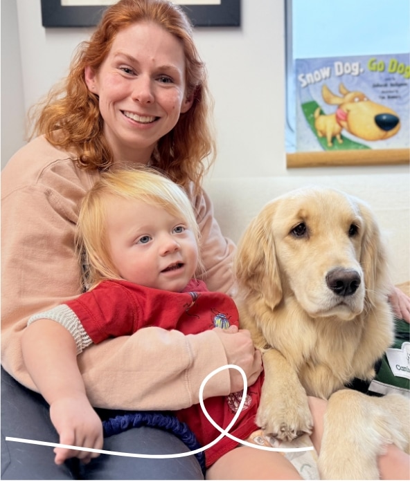 A smiling woman sits on a couch holding a young child and a golden retriever. The child wears a red shirt, and a book titled "Snow Dog, Go Dog" is visible on the windowsill behind them.