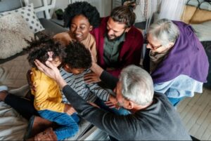 A diverse family group sits together on a bed, smiling and embracing two young children. The scene is warm and joyful, reminiscent of the togetherness often found at community events like the Kimble Tile Golf Outing, with adults and kids interacting closely in a cozy room.