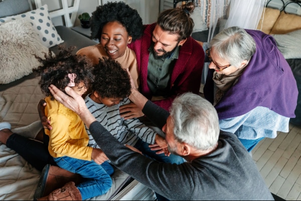 A diverse family group sits together on a bed, smiling and embracing two young children. The scene is warm and joyful, reminiscent of the togetherness often found at community events like the Kimble Tile Golf Outing, with adults and kids interacting closely in a cozy room.