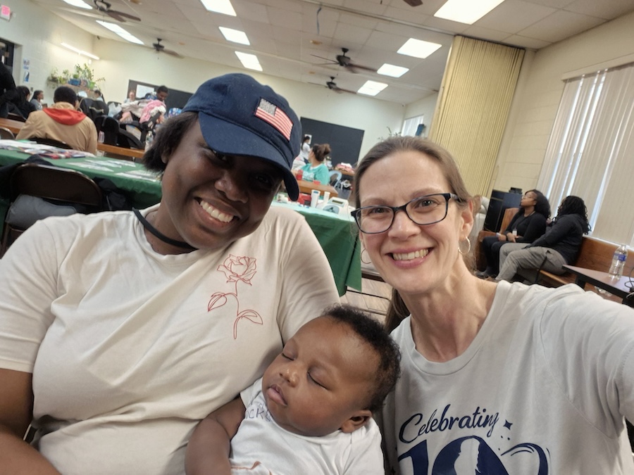 Two women smile at the camera while sitting indoors. One holds a sleeping baby. The room is filled with people seated at tables. The atmosphere appears friendly and relaxed.