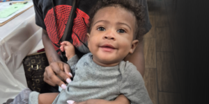 A smiling baby with curly hair sits on an adult’s lap, wearing a gray outfit. The adult, mostly out of frame, holds the baby and is dressed in a black and red shirt with pink nails visible.