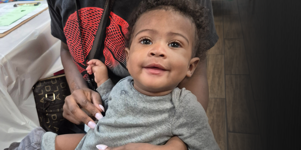 A smiling baby with curly hair sits on an adult’s lap, wearing a gray outfit. The adult, mostly out of frame, holds the baby and is dressed in a black and red shirt with pink nails visible.