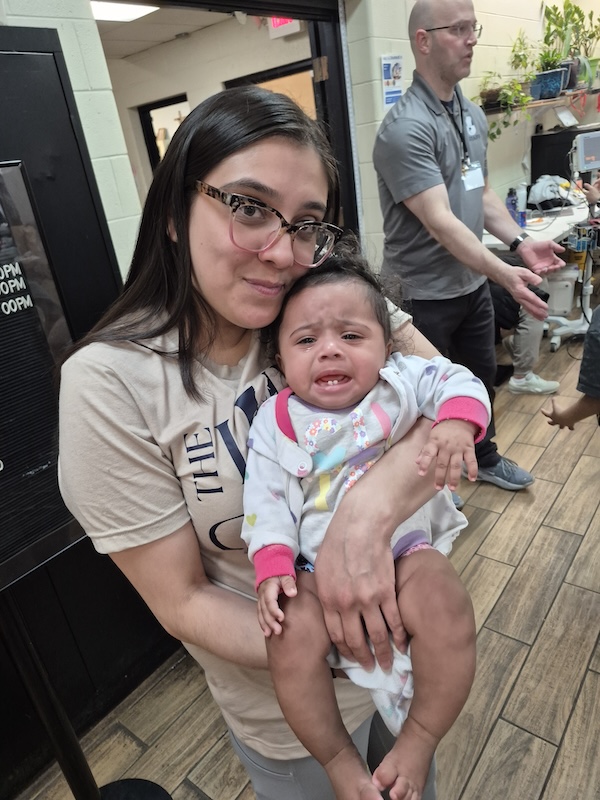 A woman with glasses holds a crying baby in a busy indoor setting. Other people and medical equipment are visible in the background.